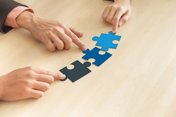 Tre mani che uniscono tessere di un puzzle su un tavolo di legno, a simboleggiare collaborazione, strategia e costruzione condivisa.