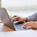 Persona che lavora al computer portatile su una scrivania, con una matita in mano e documenti appoggiati.