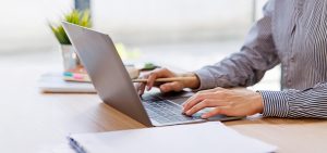 Persona che lavora al computer portatile su una scrivania, con una matita in mano e documenti appoggiati.