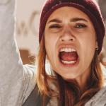 Primo piano di una donna che urla con il pugno alzato durante una manifestazione per i diritti. La donna indossa un berretto e una felpa, e sullo sfondo si intravedono dei cartelli.