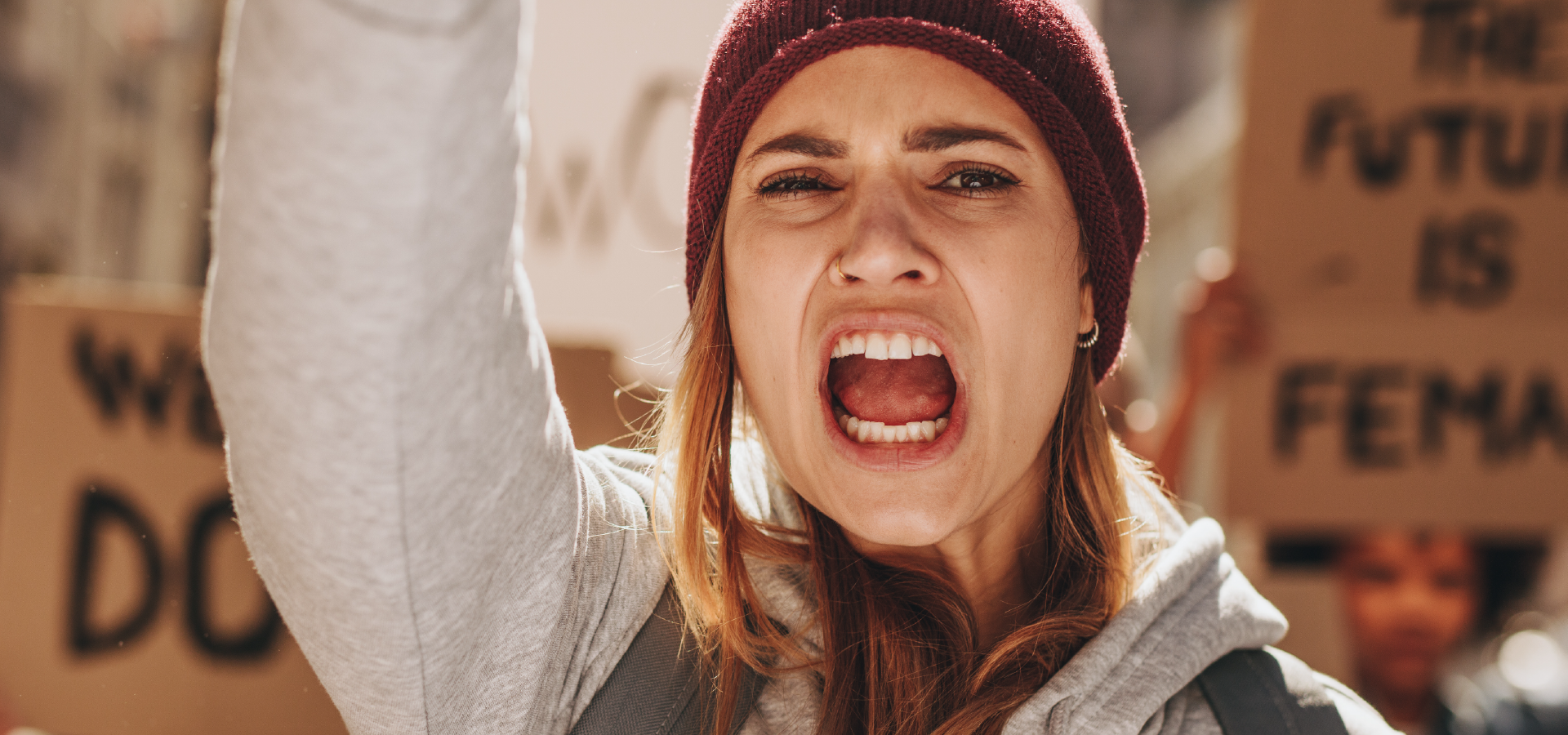 Primo piano di una donna che urla con il pugno alzato durante una manifestazione per i diritti. La donna indossa un berretto e una felpa, e sullo sfondo si intravedono dei cartelli.