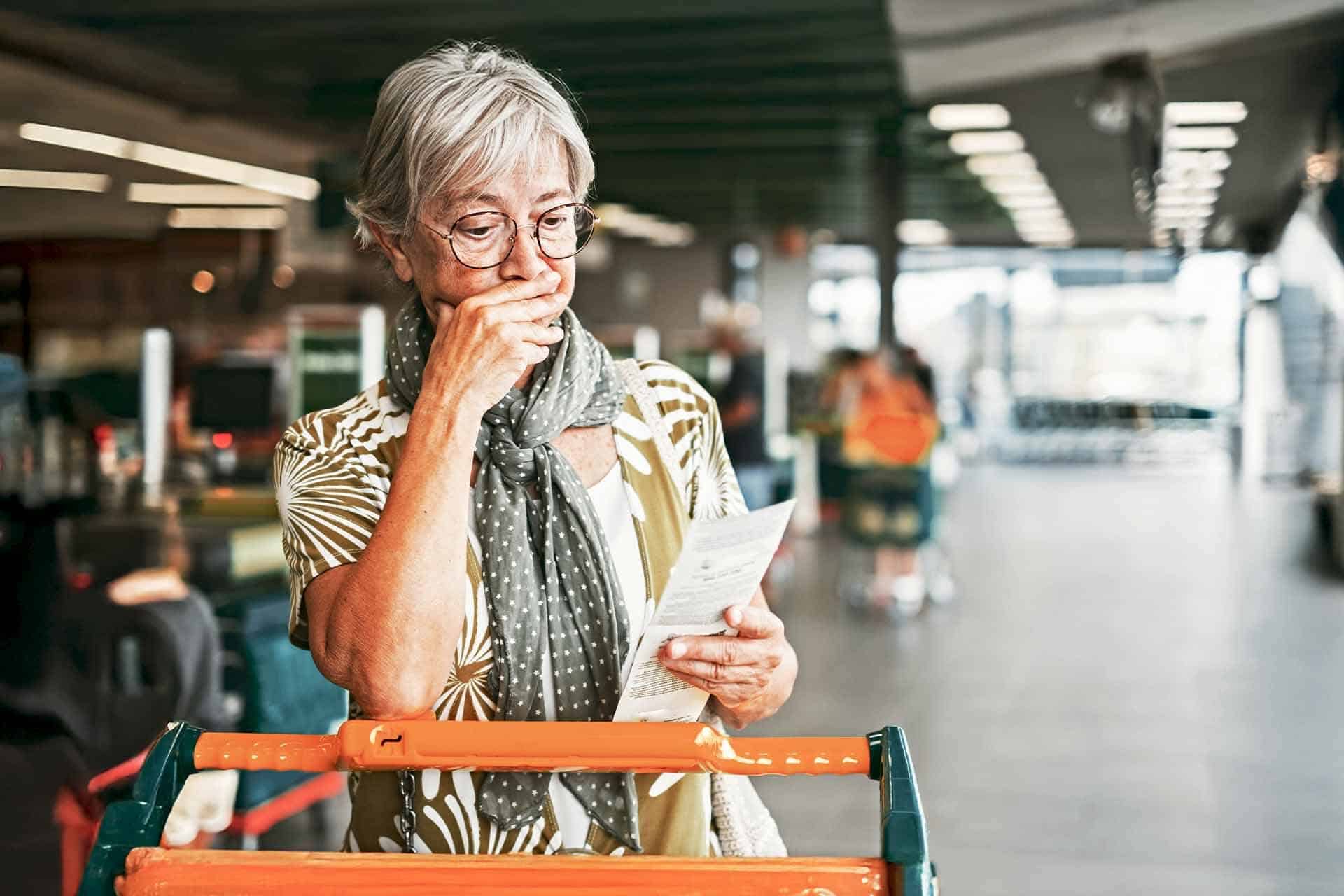 Una donna anziana in un supermercato, con espressione preoccupata, tiene in mano una ricevuta, simboleggiando l'impatto dell'inflazione sulla vita quotidiana.