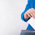 Una persona che indossa una giacca blu inserisce una busta bianca elettorale, riportante il logo del Fondo Telemaco, all'interno di un'urna.