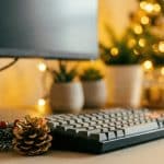Dettaglio di una scrivania d'ufficio con tastiera e mouse, decorata con una pigna e un rametto natalizio in primo piano; sullo sfondo sfocato si vedono luci di Natale calde e luminose che creano un'atmosfera festiva.