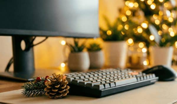 Dettaglio di una scrivania d'ufficio con tastiera e mouse, decorata con una pigna e un rametto natalizio in primo piano; sullo sfondo sfocato si vedono luci di Natale calde e luminose che creano un'atmosfera festiva.