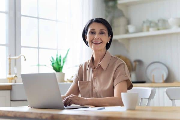 Donna sorridente seduta in cucina che utilizza un laptop per informarsi sulla pensione anticipata 2026.