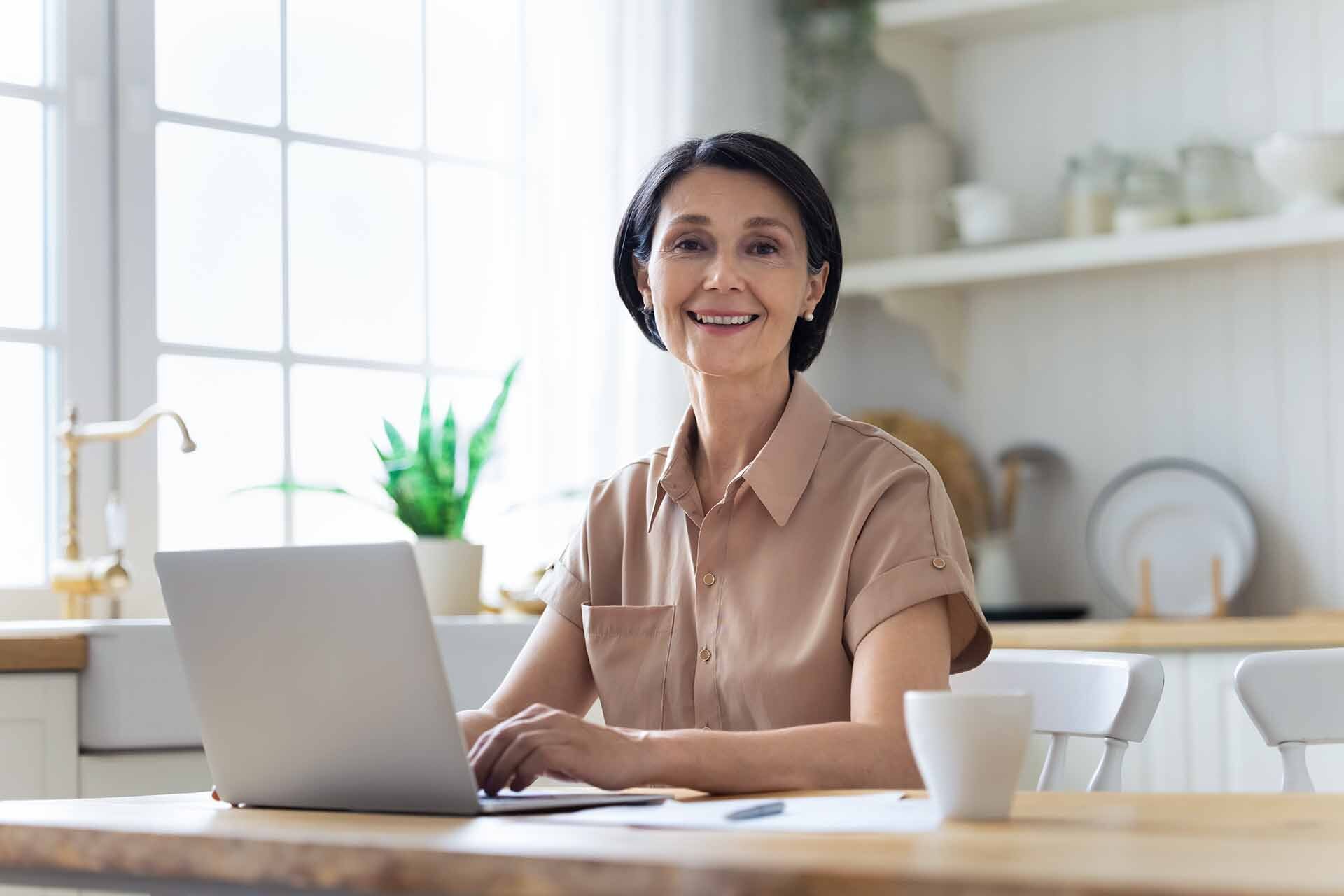 Donna sorridente seduta in cucina che utilizza un laptop per informarsi sulla pensione anticipata 2026.