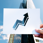 Una mano tiene in primo piano un cartoncino bianco con una freccia rivolta verso l'alto ritagliata al centro. Sullo sfondo si stagliano moderni grattacieli specchiati sotto un limpido cielo azzurro, simboleggiando crescita e performance positive.