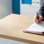 Un professionista in giacca scura e camicia bianca è ritratto mentre firma una pila di documenti appoggiati su una scrivania in legno chiaro. L'azione avviene all'interno di un ufficio luminoso; la mano destra impugna una penna stilografica nera con finiture dorate, mentre la sinistra tiene fermo il plico inserito in una cartella in pelle.