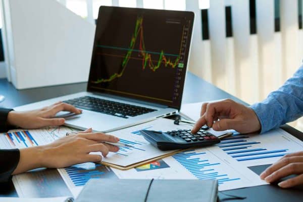 Analisi tecnica del rischio valutario: professionisti confrontano grafici di trading su laptop e report cartacei con l'ausilio di una calcolatrice.