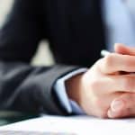 Primo piano delle mani incrociate di un professionista in abito scuro che tiene una penna, appoggiate su documenti aziendali