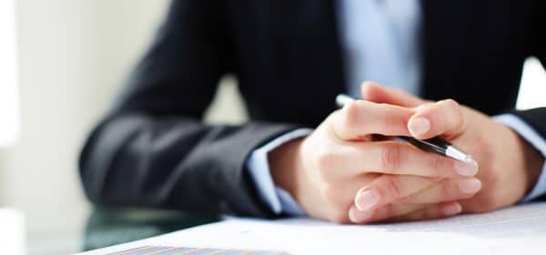 Primo piano delle mani incrociate di un professionista in abito scuro che tiene una penna, appoggiate su documenti aziendali