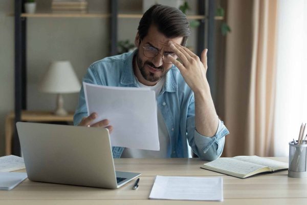 Un uomo preoccupato analizza documenti finanziari al computer, rappresentando l'ansia finanziaria e l'avversione al rischio riguardo alla pensione futura.