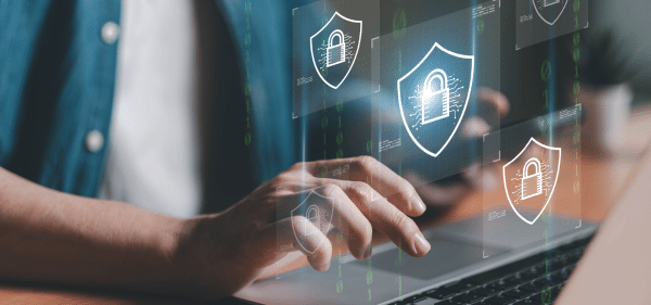 Un primo piano delle mani di una persona che digita su un computer portatile, con icone olografiche fluttuanti di scudi di sicurezza digitali contenenti lucchetti, e flussi di codice binario sfocato sullo sfondo.