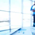 In primo piano su un tavolo di vetro, documenti con grafici aziendali colorati, una penna e un tablet sono a fuoco. Sullo sfondo sfuocato, un uomo d'affari in camicia blu è in piedi e parla al telefono in un ufficio moderno e luminoso con finestre a tutta altezza.