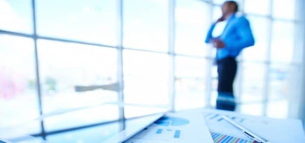 In primo piano su un tavolo di vetro, documenti con grafici aziendali colorati, una penna e un tablet sono a fuoco. Sullo sfondo sfuocato, un uomo d'affari in camicia blu è in piedi e parla al telefono in un ufficio moderno e luminoso con finestre a tutta altezza.