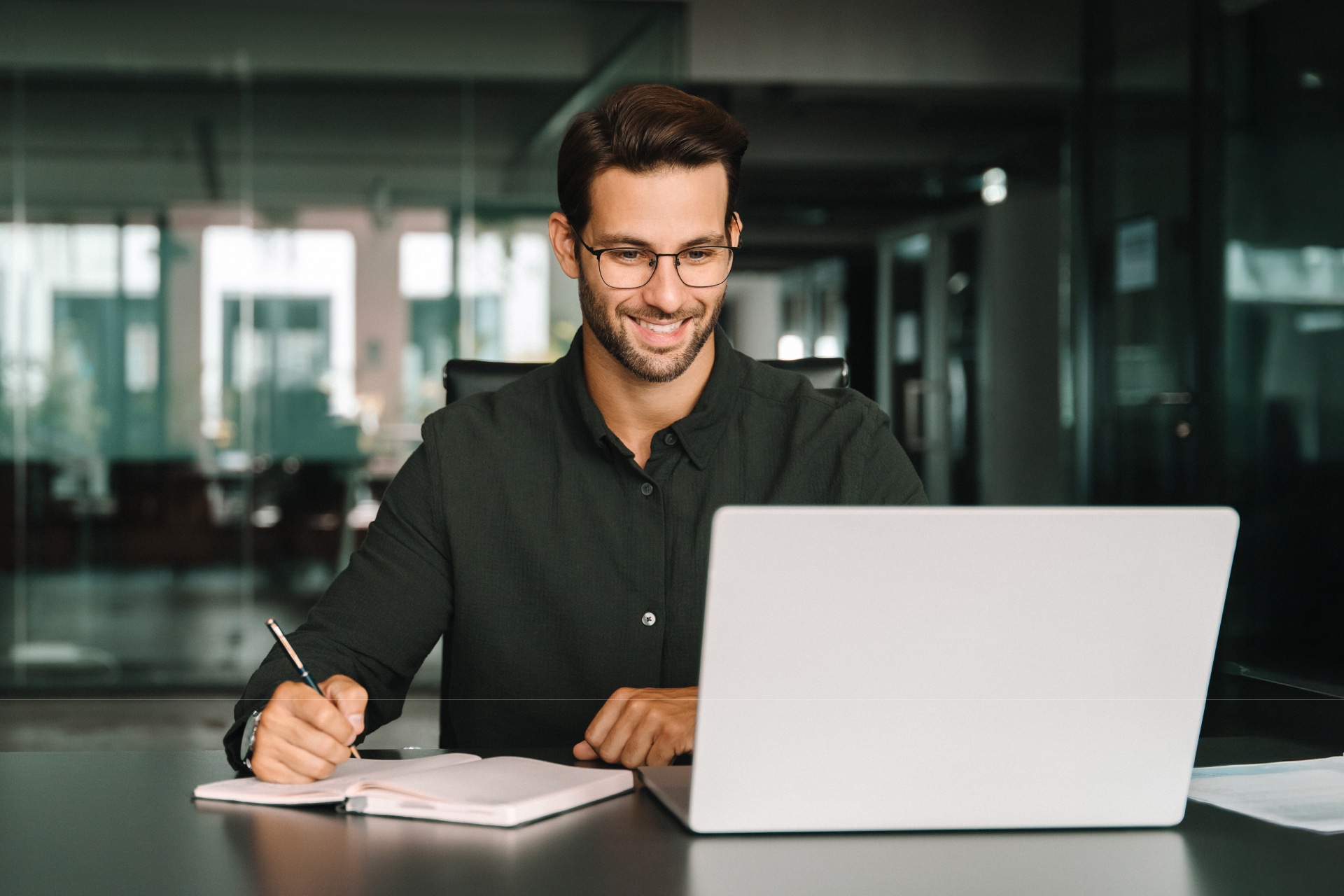 Professionista sorridente in ufficio che prende appunti su un'agenda mentre analizza dati finanziari al computer portatile.