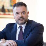 Foto ufficiale di Stefano Castrignanò, nuovo Presidente di Fondo Telemaco, seduto a una scrivania in un ufficio, con abito blu e cravatta bordeaux, mentre guarda verso l’obiettivo.