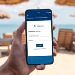 Una mano tiene uno smartphone che visualizza la schermata di login dell'App del Fondo Telemaco. Lo sfondo è una spiaggia sfocata con ombrelloni e il mare.