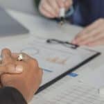 Dettaglio delle mani di due professionisti durante una riunione, intenti ad analizzare documenti finanziari, grafici e fogli di calcolo su una scrivania con una calcolatrice bianca e un computer portatile.