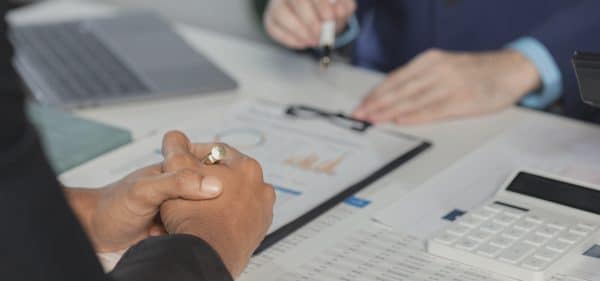 Dettaglio delle mani di due professionisti durante una riunione, intenti ad analizzare documenti finanziari, grafici e fogli di calcolo su una scrivania con una calcolatrice bianca e un computer portatile.